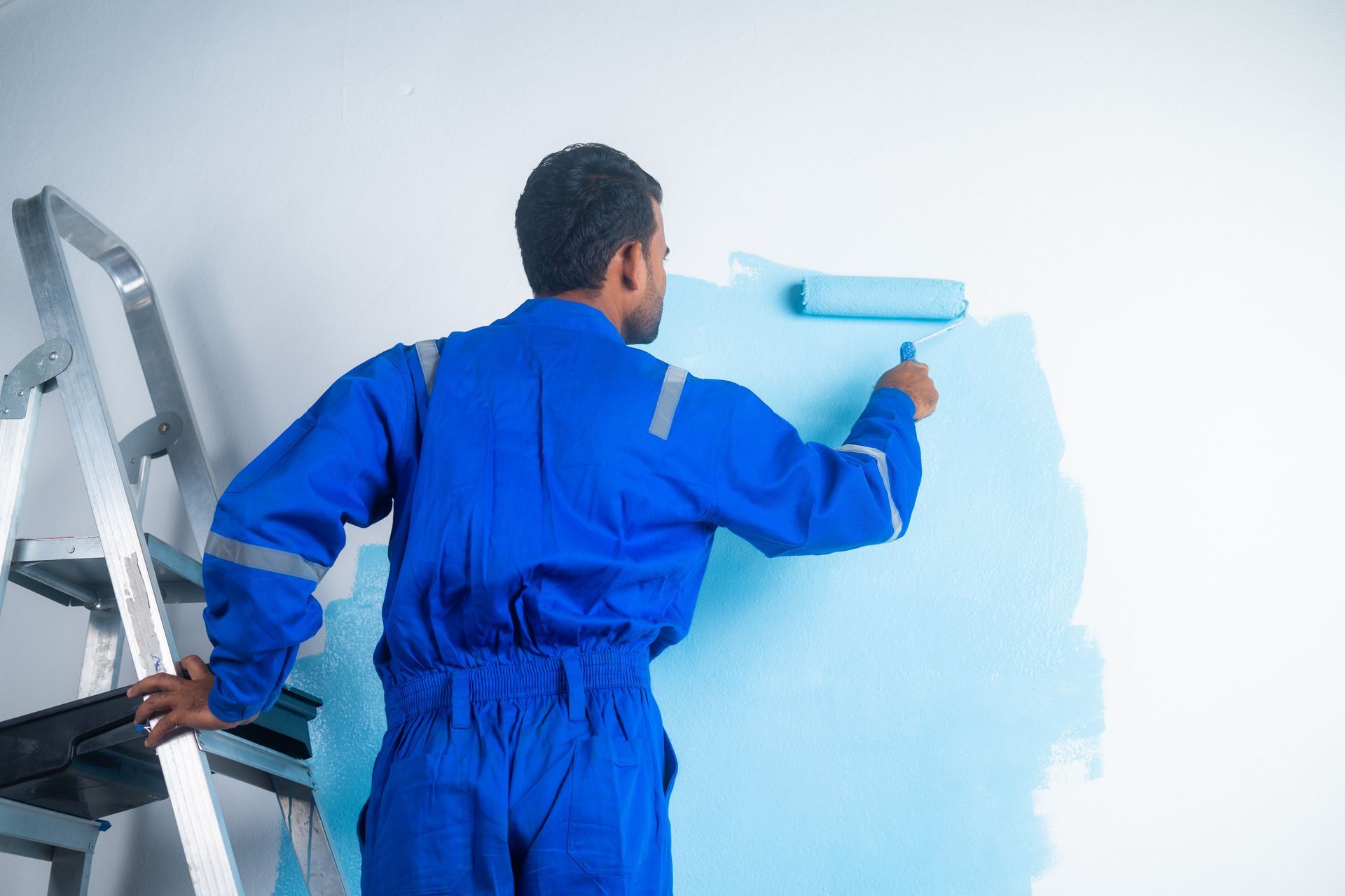 A man in a blue jumpsuit is painting a wall with a roller.