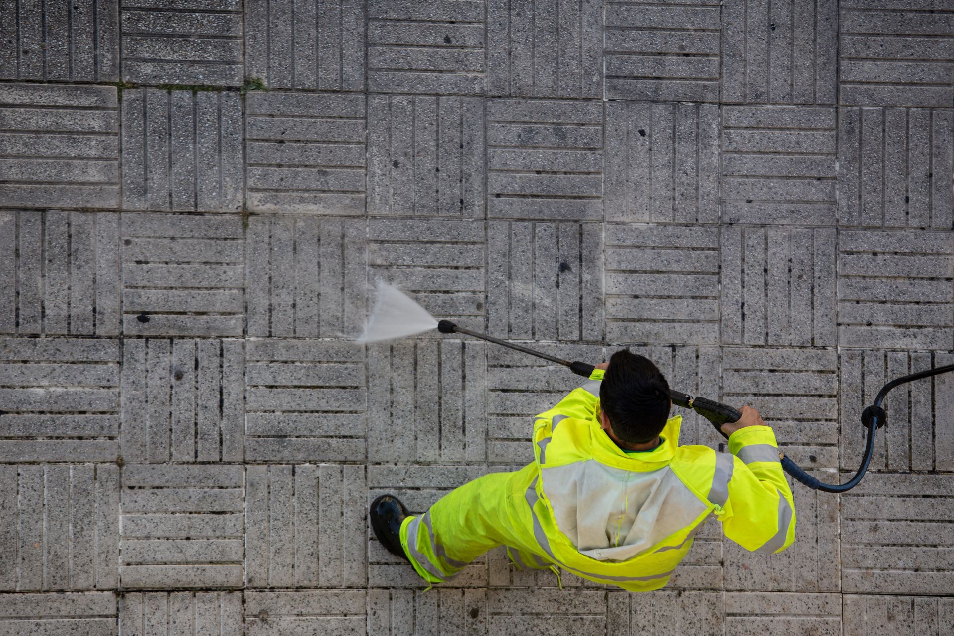 A man in a yellow jacket is using a high pressure washer to clean a tiled floor.