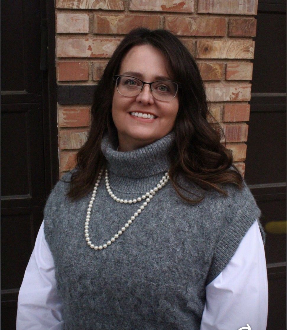 Woman with glasses, wearing a pearl necklace, gray sweater, and white shirt. Standing in front of brick wall.