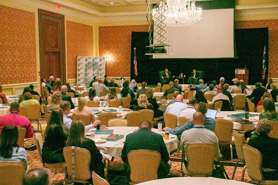 A conference room with attendees seated at round tables facing a stage with speakers and a screen.