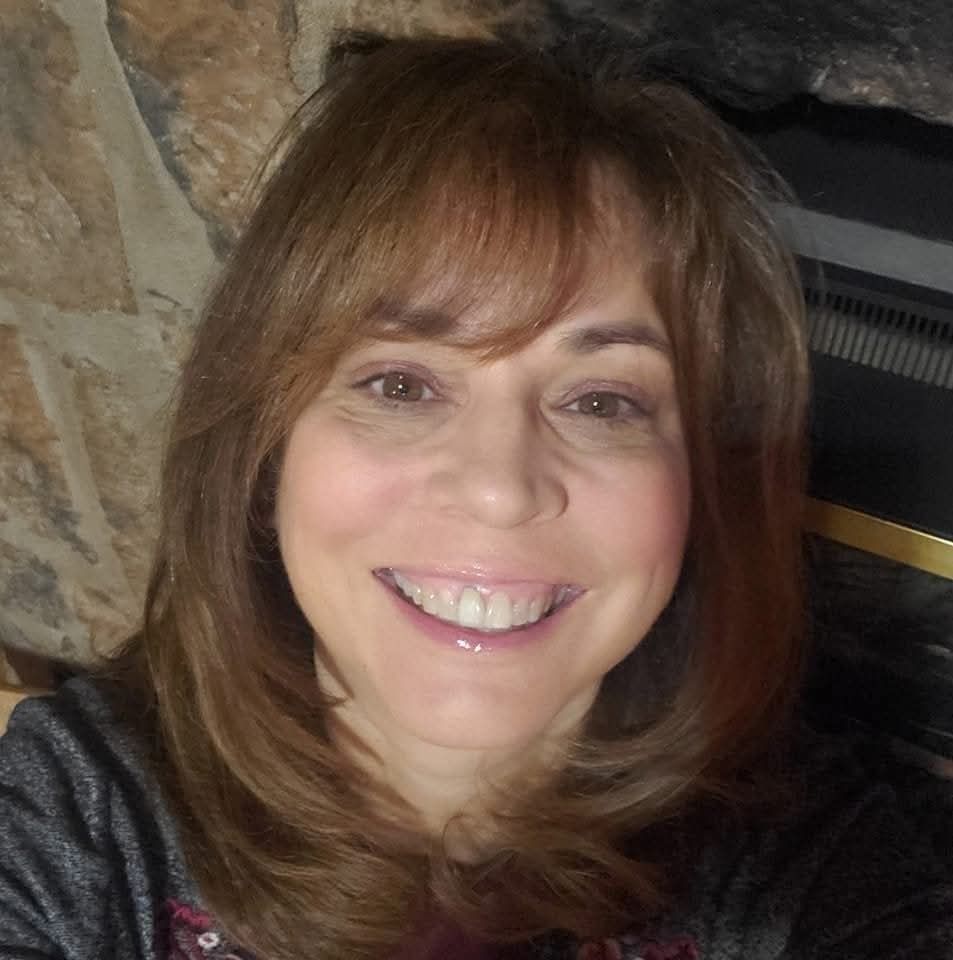 Woman with shoulder-length brown hair smiling in front of a stone wall and fireplace.