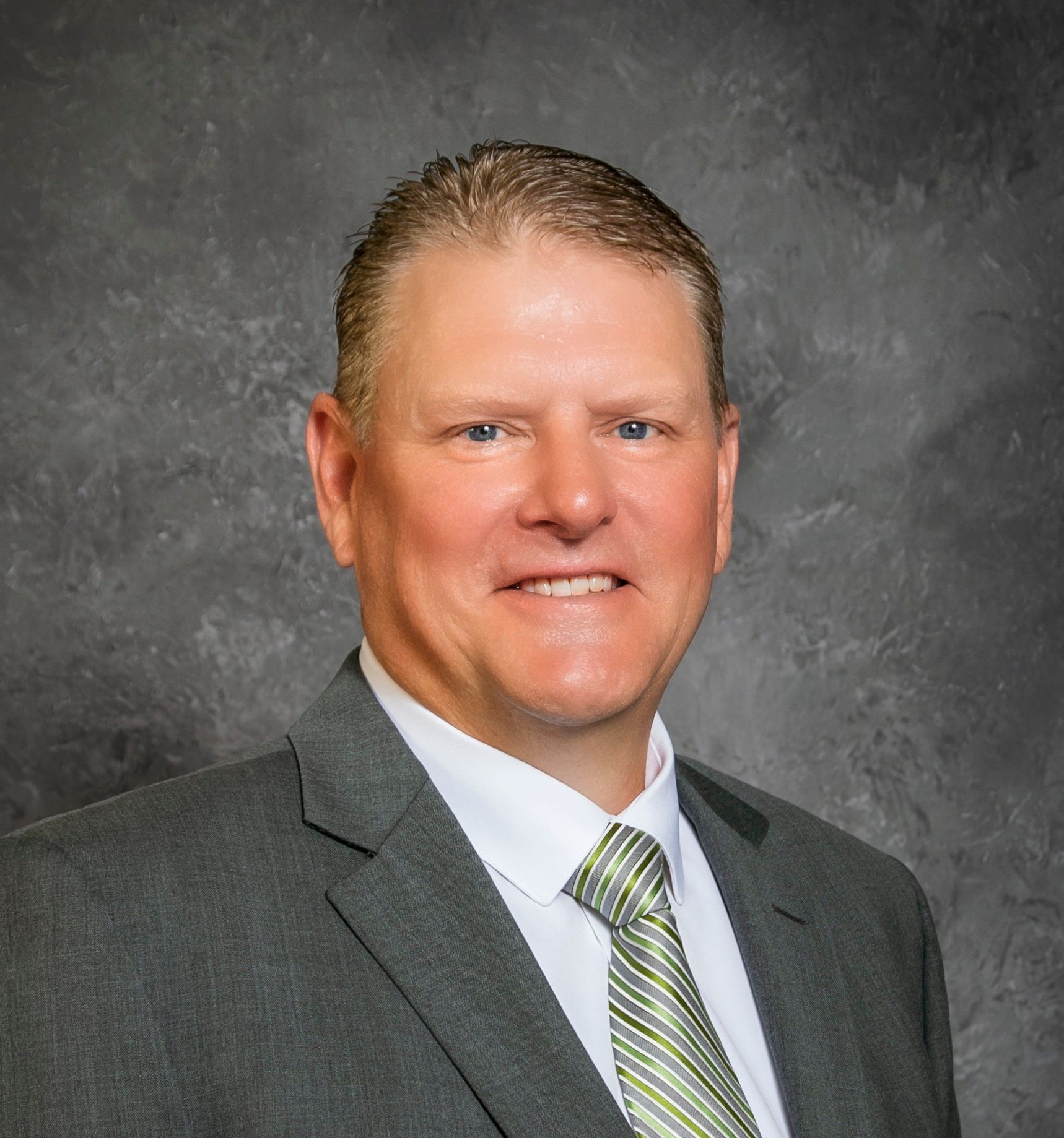 Man in a gray suit and green patterned tie, smiling against a mottled gray background.