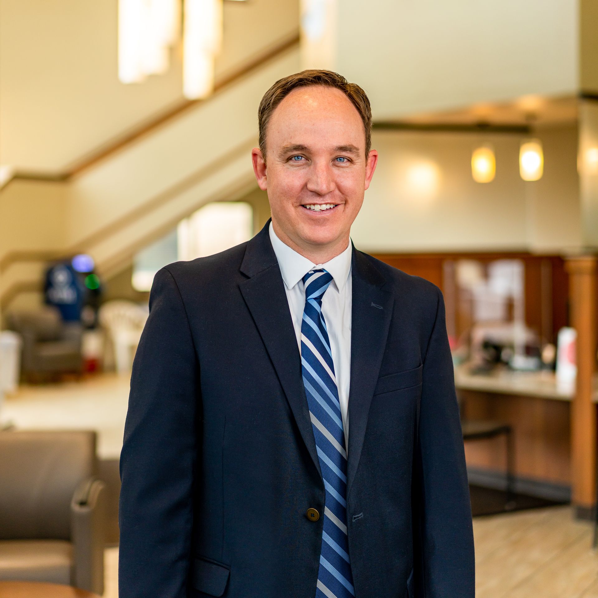 Man in a navy suit and striped tie smiles in an office lobby with a staircase.
