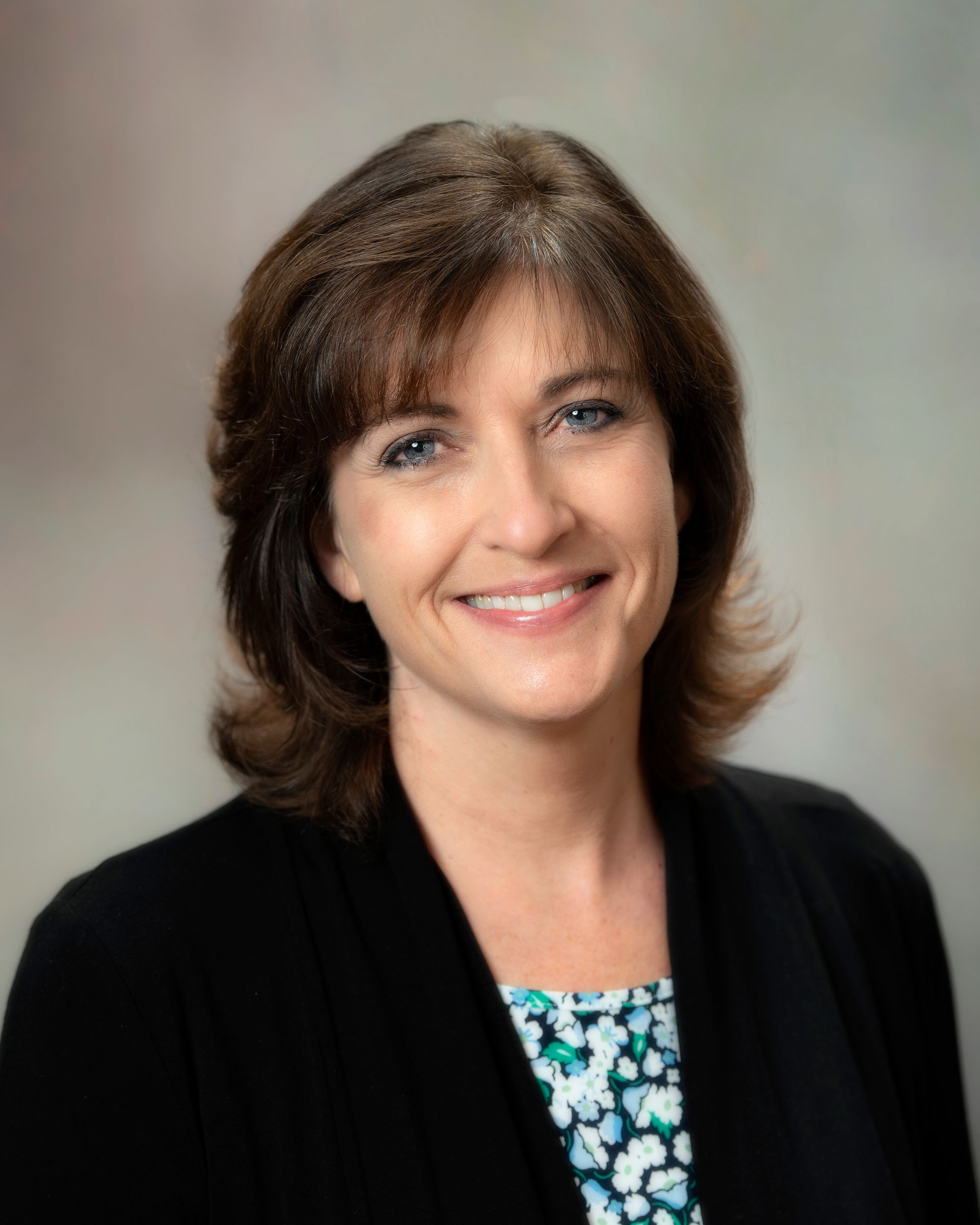 Woman with dark hair smiles, wearing a black blazer over a patterned top, against a soft background.