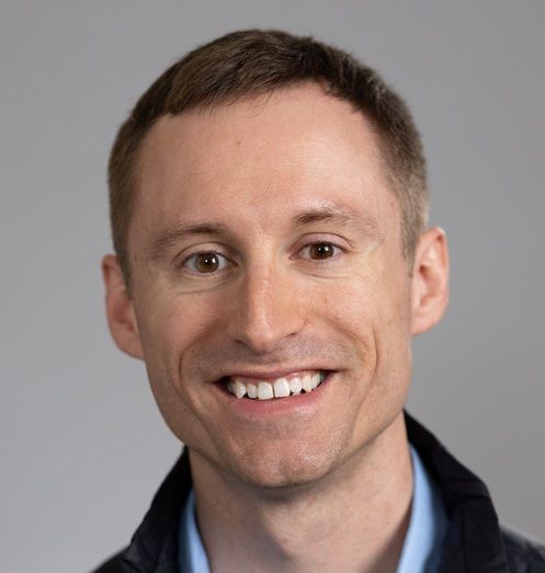 Man in a light blue shirt and dark jacket smiles at the camera.