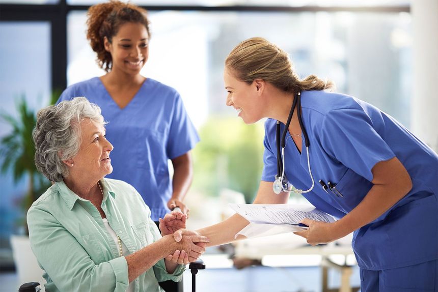 Two medical professionals in blue scrubs shake hands with a smiling patient in a wheelchair.