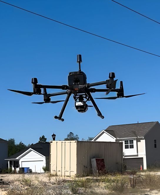 A black, multi-rotor industrial drone hovers in a clear blue sky above a residential construction site.