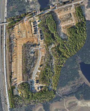 An aerial view of a construction site with cleared earth, partially built residential structures, and surrounding woods.