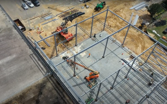 Aerial view of a metal building frame under construction with heavy machinery on a dirt lot.