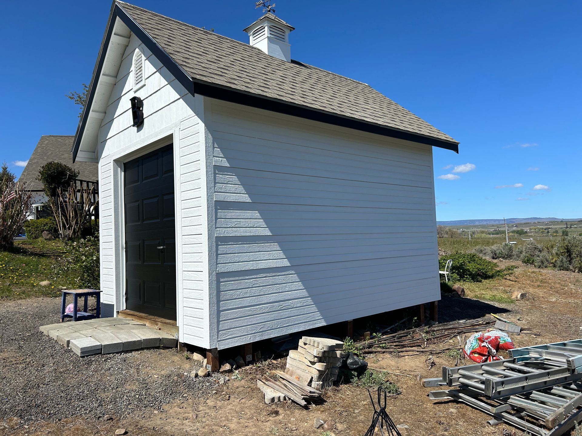 A small white shed with a black door is in the middle of a dirt field.