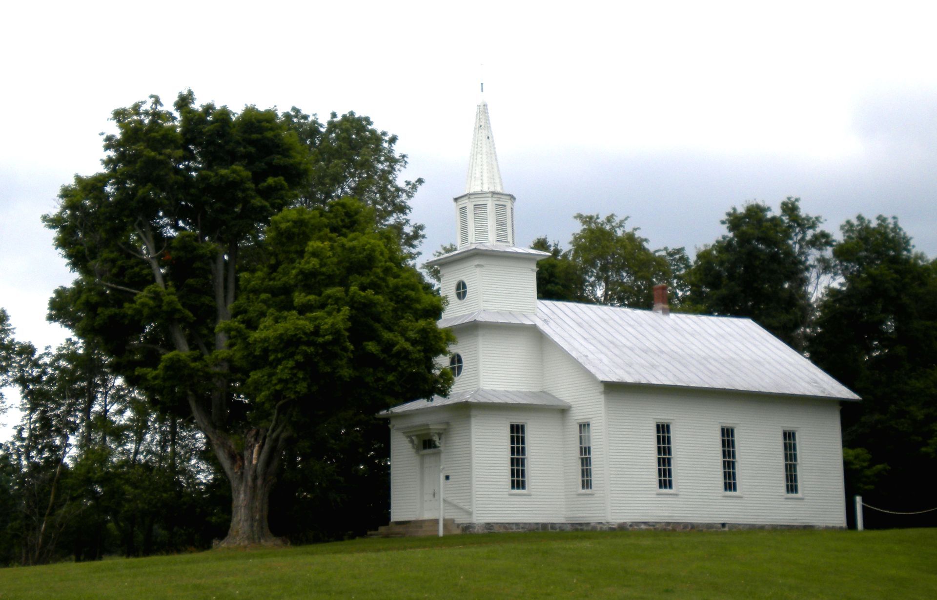White church with steeple on a grassy hill, surrounded by trees under a cloudy sky