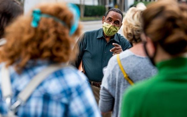 Masked speaker addressing a small crowd outdoors, with people listening in the foreground.