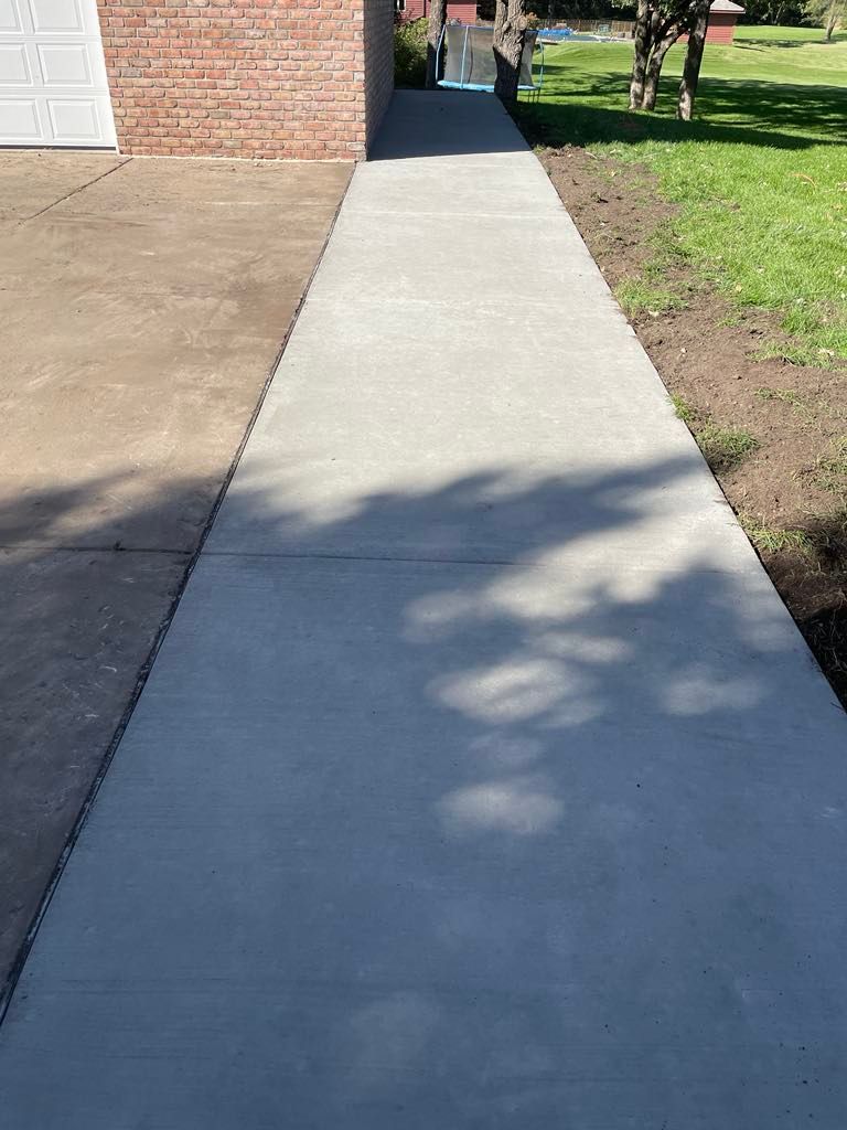 A concrete walkway leading to a house with a brick building in the background.