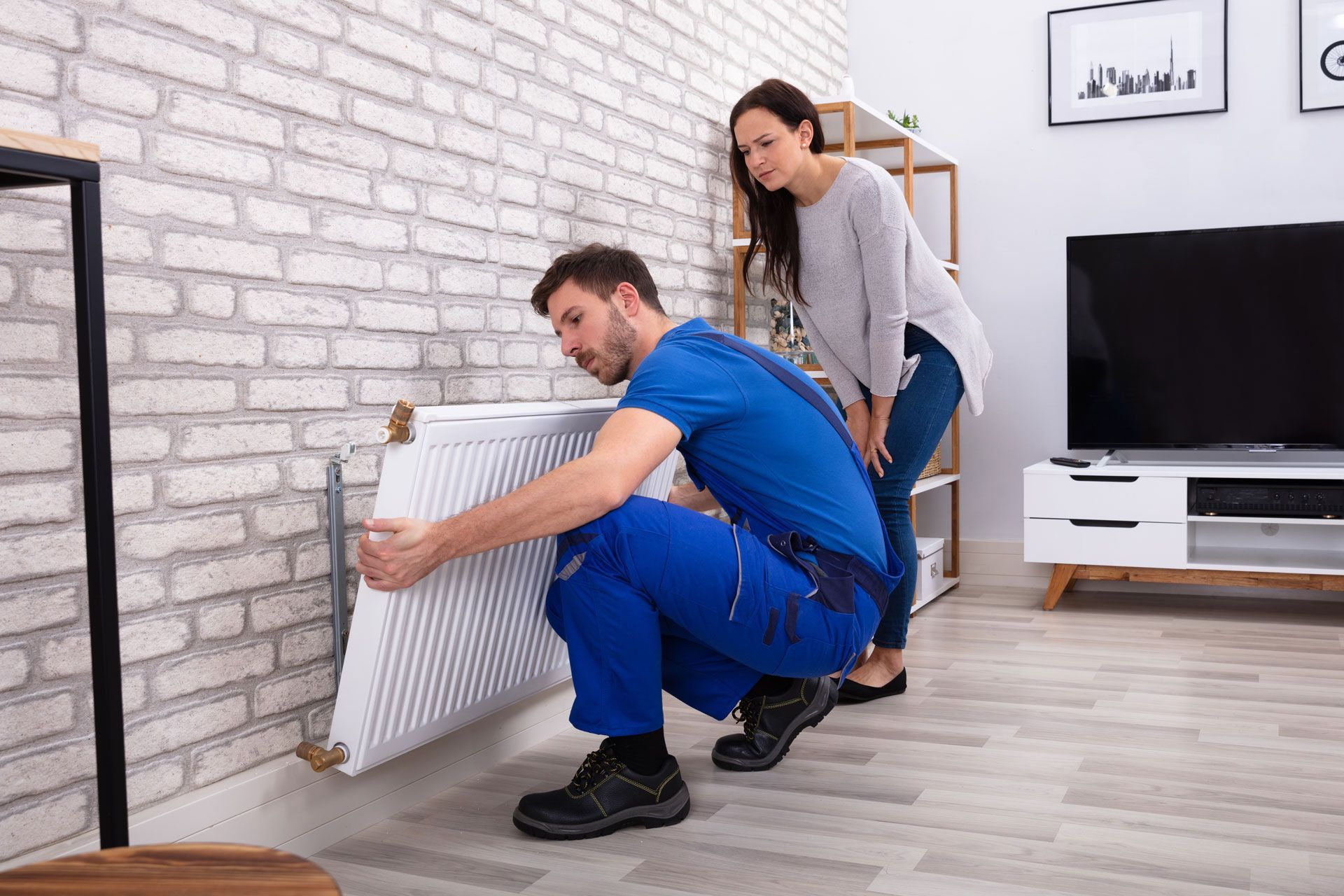 A man is installing a radiator in a living room while a woman watches.