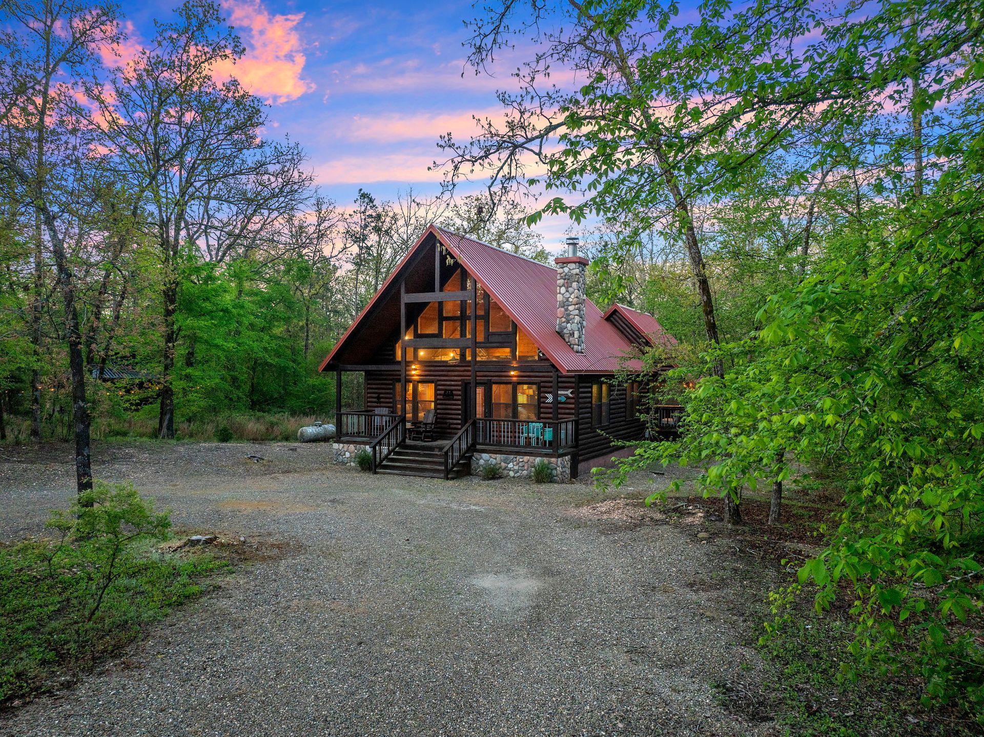 A rustic A-frame cabin with a red metal roof sits among lush green trees at sunset with a gravel driveway in front.