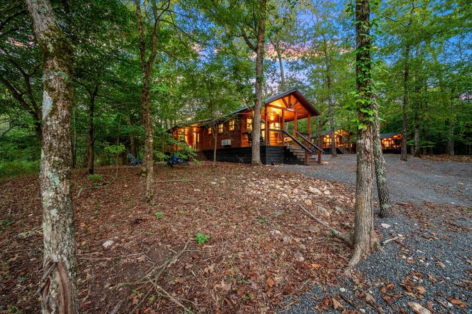 A warm, glowing wooden cabin tucked among lush green trees, accessible by a gravel path during the golden hour.