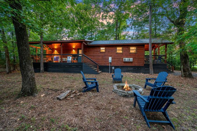 A rustic wooden cabin with a lit porch and deck, surrounded by trees, with a stone fire pit and blue chairs in front.