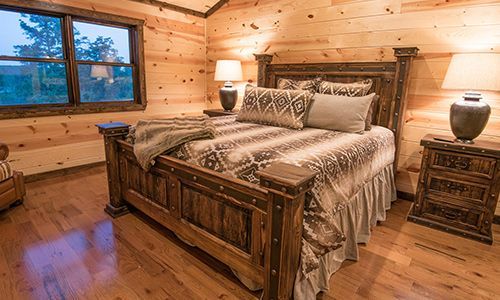 A bedroom in a log cabin with a wooden bed , nightstands , lamps and a large window.