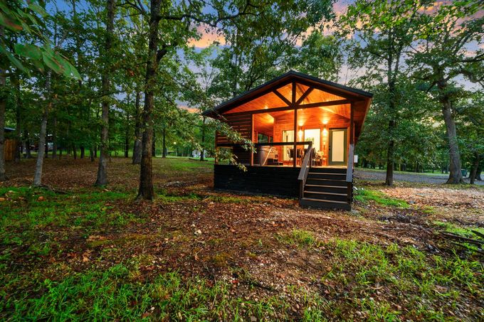 A glowing wooden cabin sits nestled in a forest of trees at sunset with fallen leaves covering the ground.