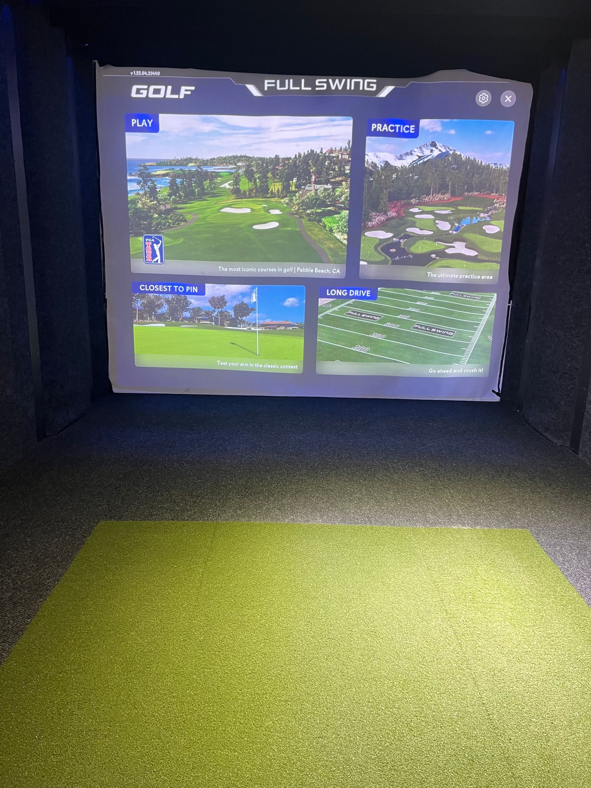 Golf simulator setup with screen displaying golf course, green turf, dark walls.