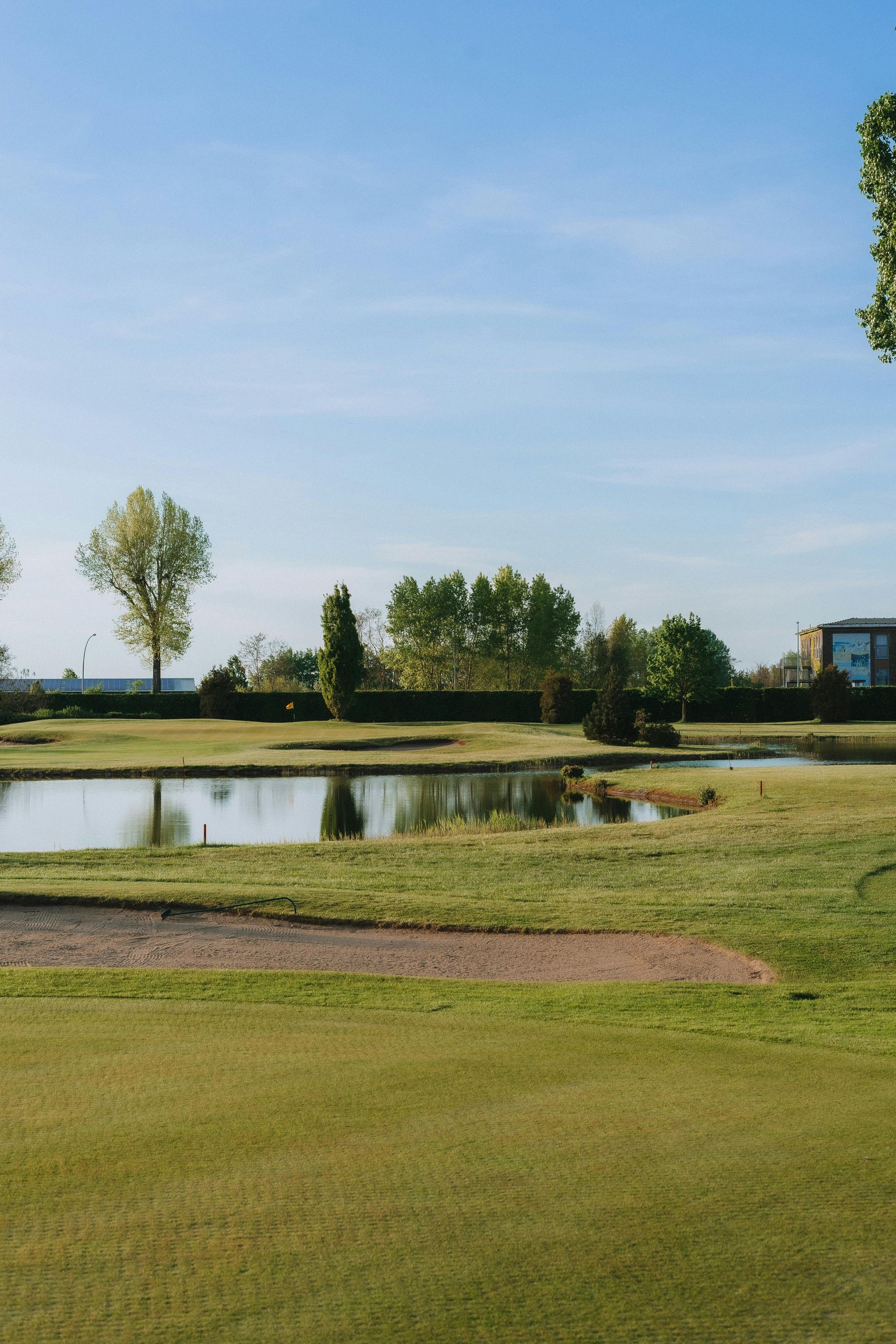 Green golf course with pond, trees, and buildings under a blue sky.