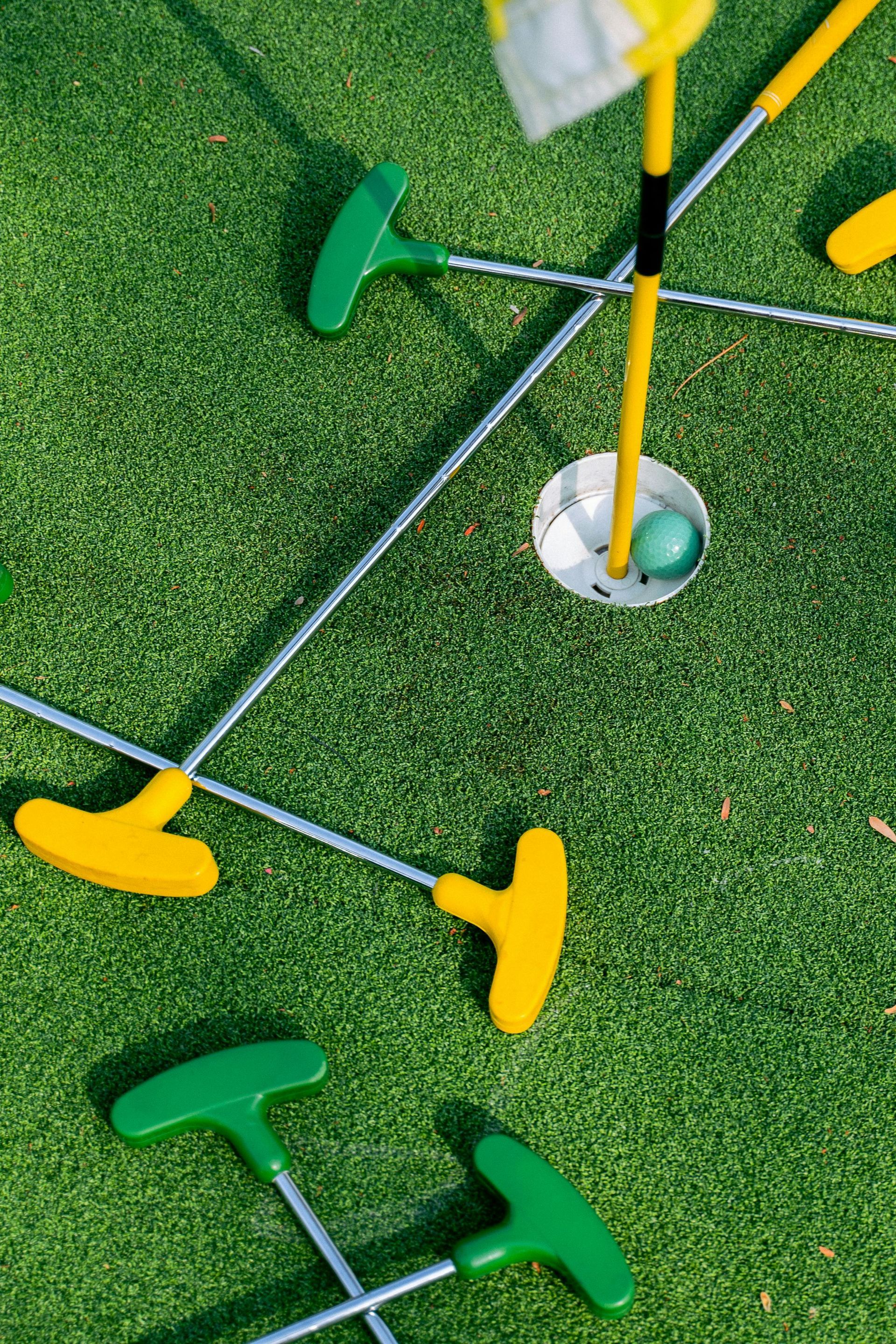 Miniature golf scene, green turf, several putters, a hole with a ball, and a yellow flag.