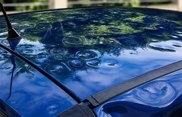 Blue car roof with circular water dents and reflections, showing a black antenna and trim strip
