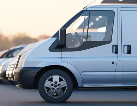 Silver delivery van driving on a road beside parked cars at dusk