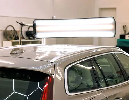 Silver car roof in a garage under bright overhead lights, with a large light fixture reflected on the paint