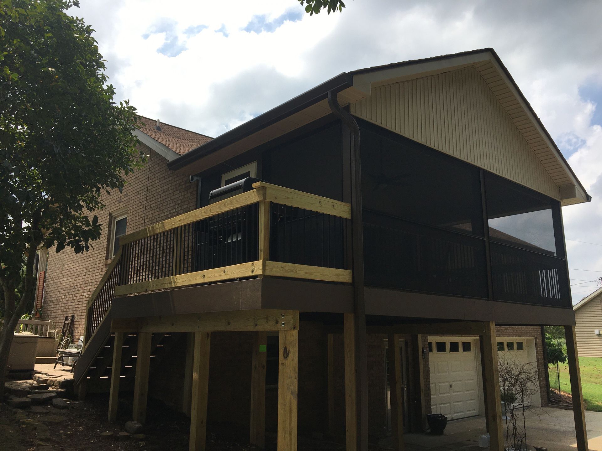 Screened porch with dark screens, brown decking, and wooden supports, next to a brick house.