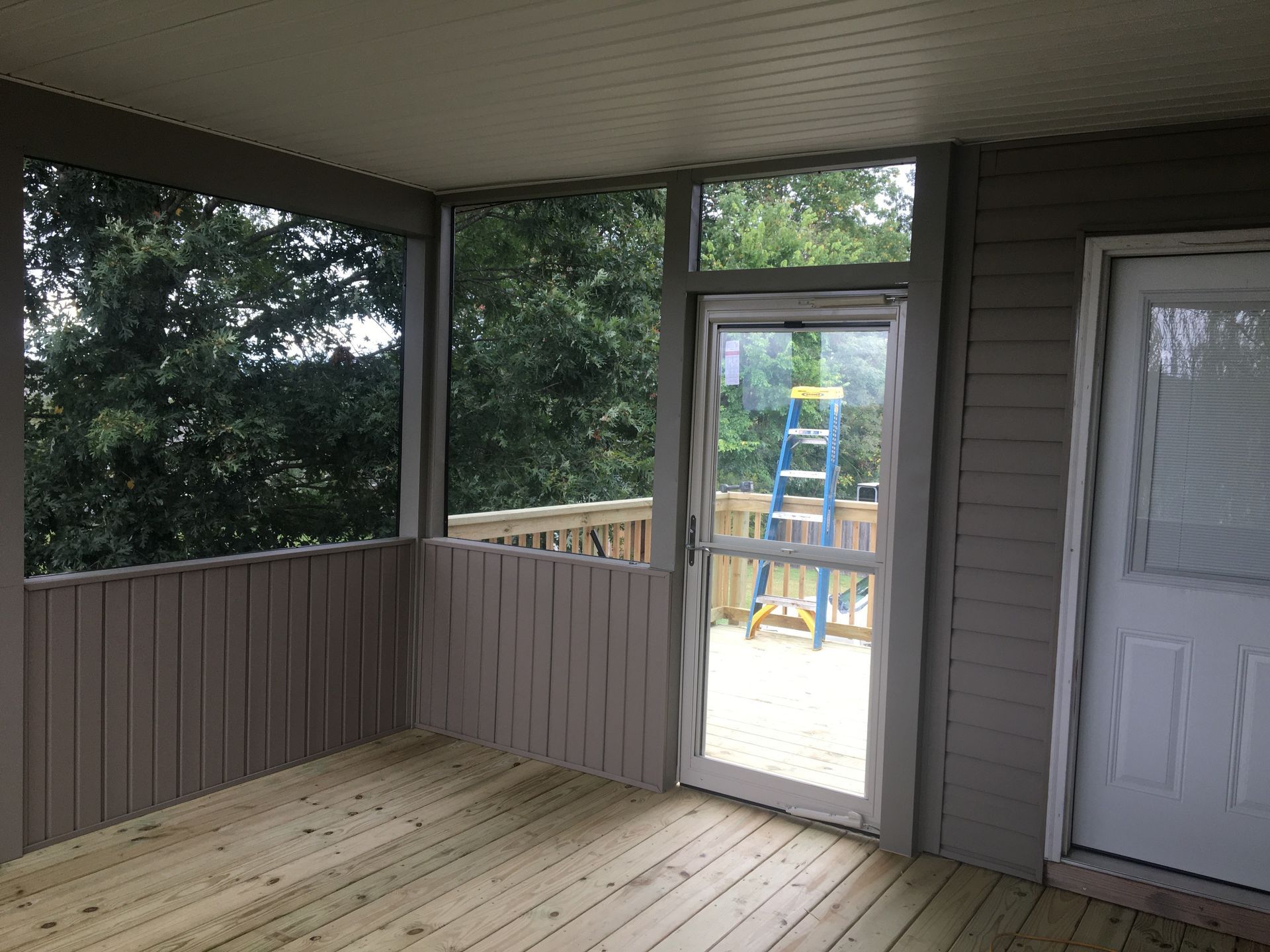 Screened-in porch with wood floor, gray walls, and glass door. Trees visible through openings.
