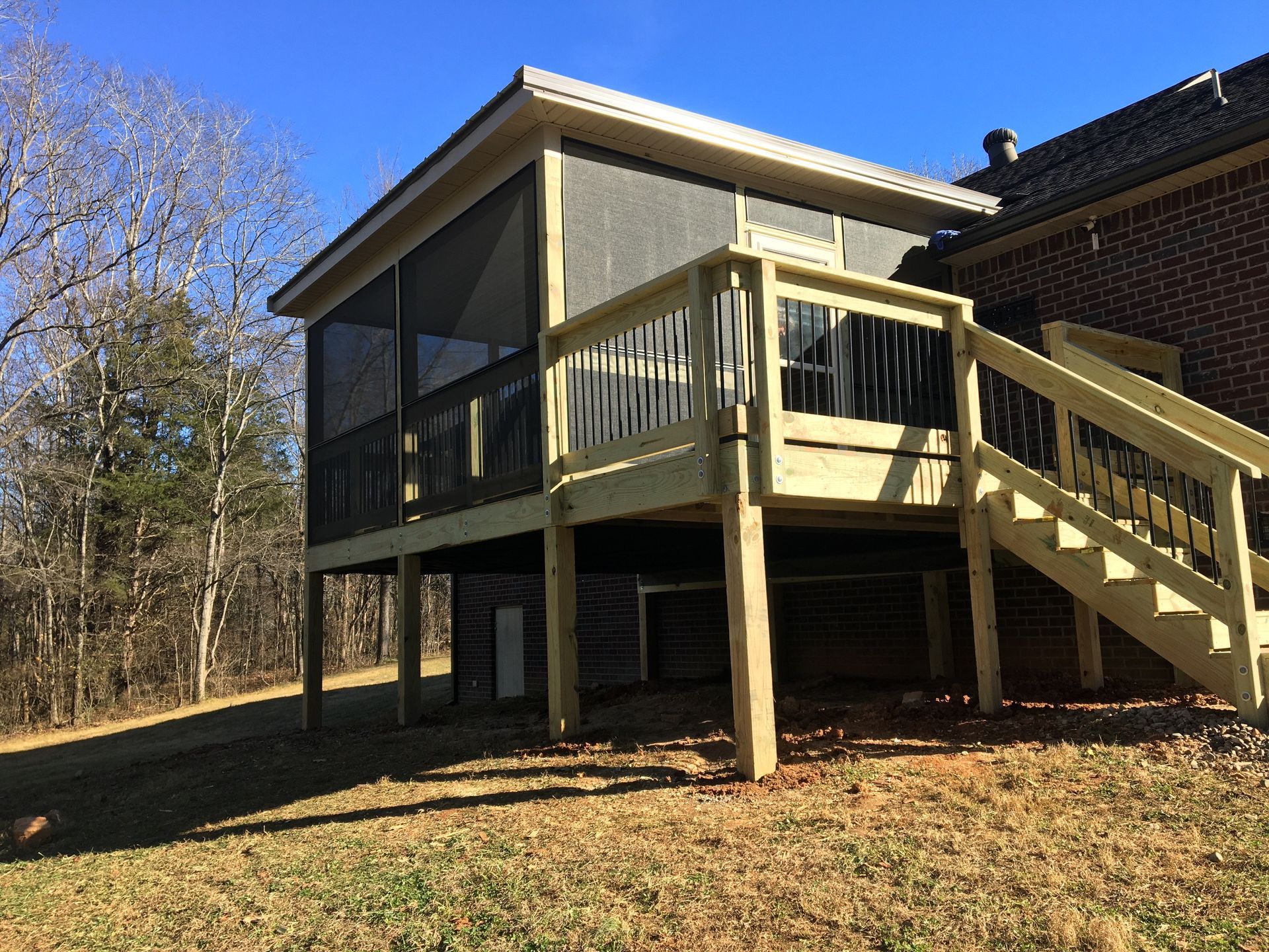 Screened porch with stairs and railing attached to a brick house on a grassy, sunny hillside.