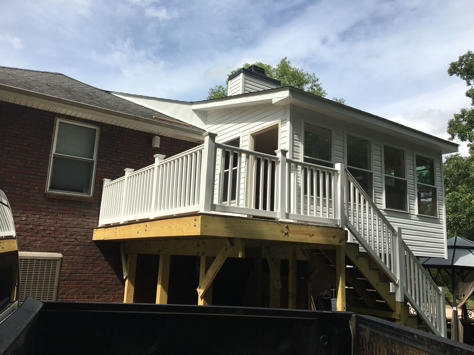 A deck with white railing attached to a brick house and an enclosed sunroom, stairs leading down.
