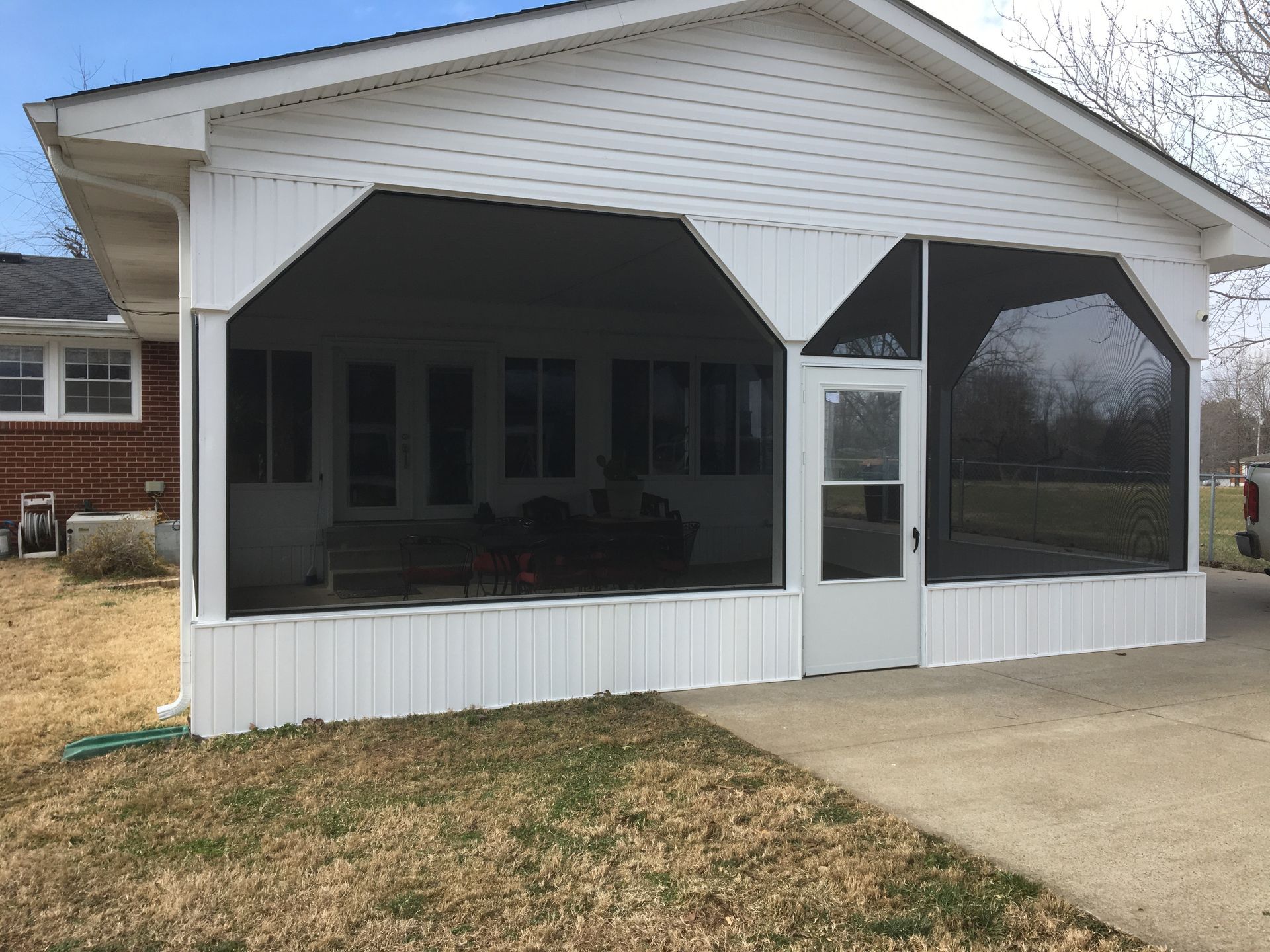White screened-in porch attached to a house; door in center, windows behind screens, driveway in front.