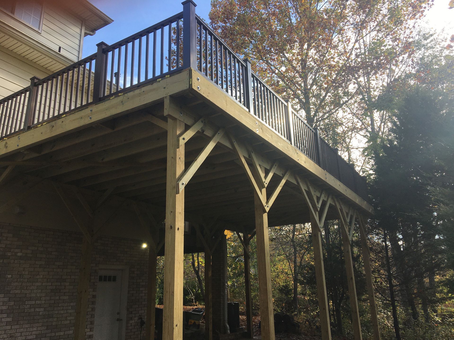 Elevated wooden deck attached to a brick house, supported by wooden posts in a wooded setting.