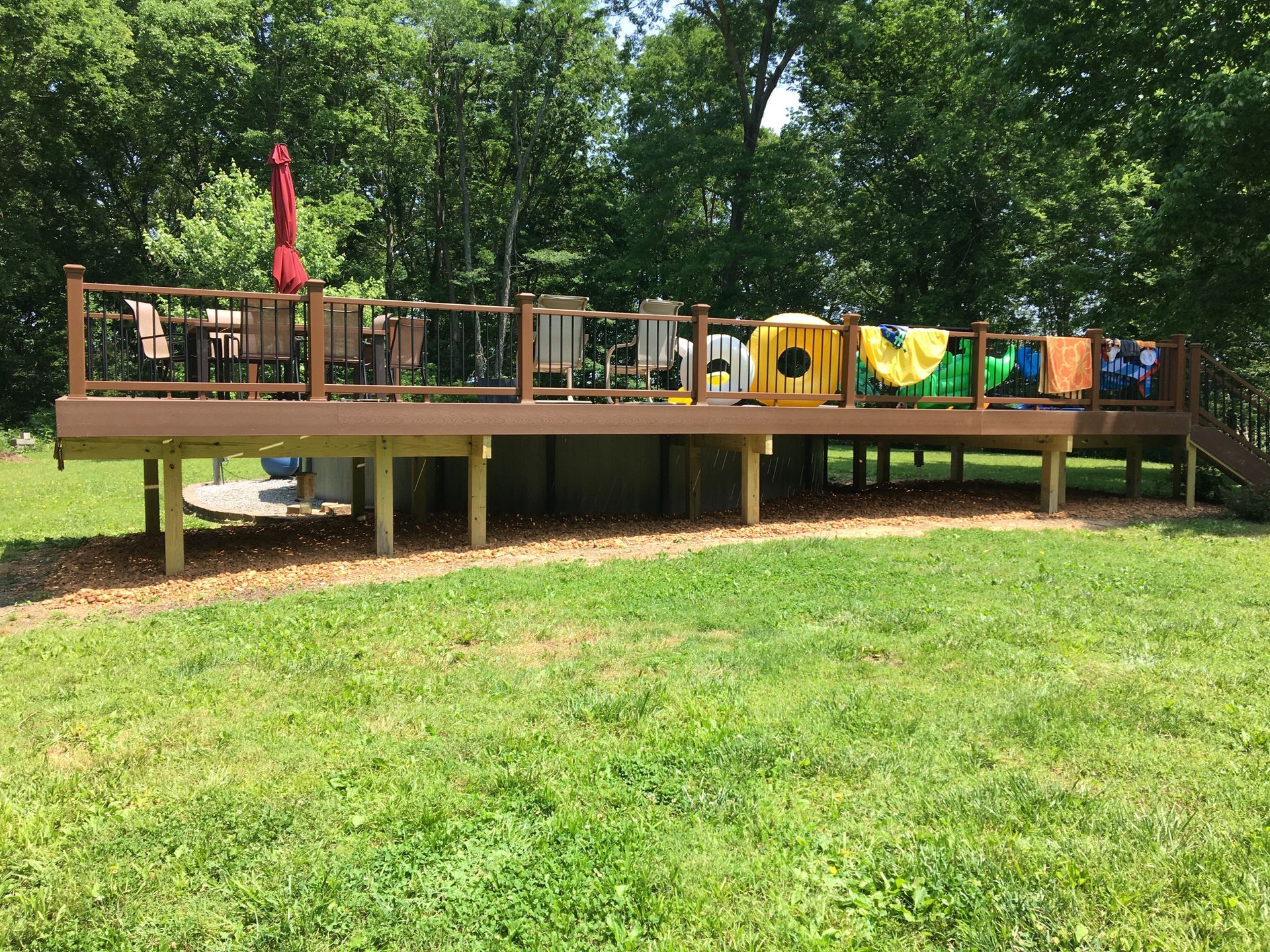 Wooden deck surrounding an above-ground pool, towels drying, red umbrella, and greenery.