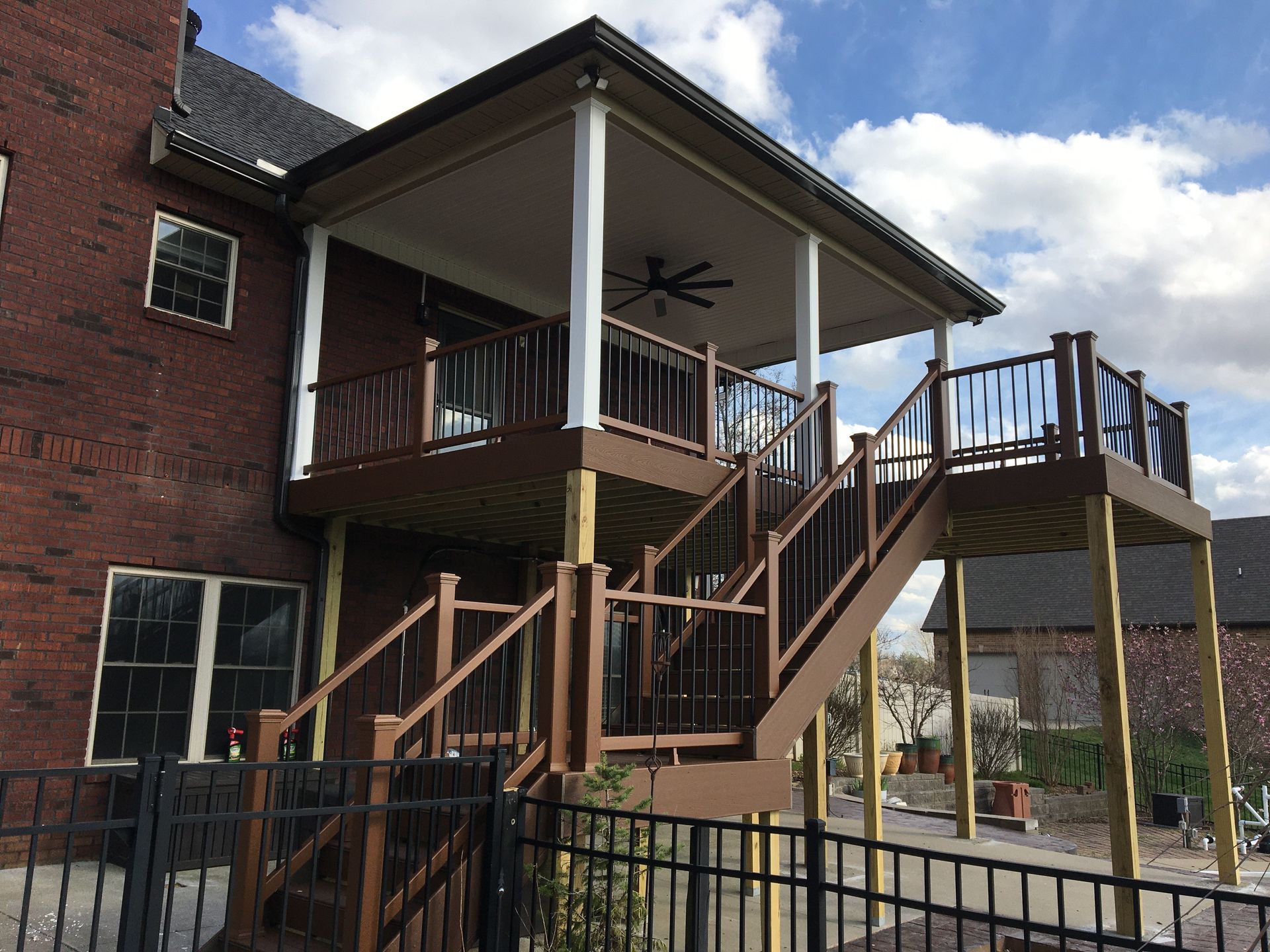 Two-story wooden deck with stairs attached to a brick house, railings are brown, the sky is blue.