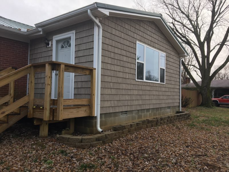 Small building with shingle siding, a deck, a window, and a door, in a yard with leaves.