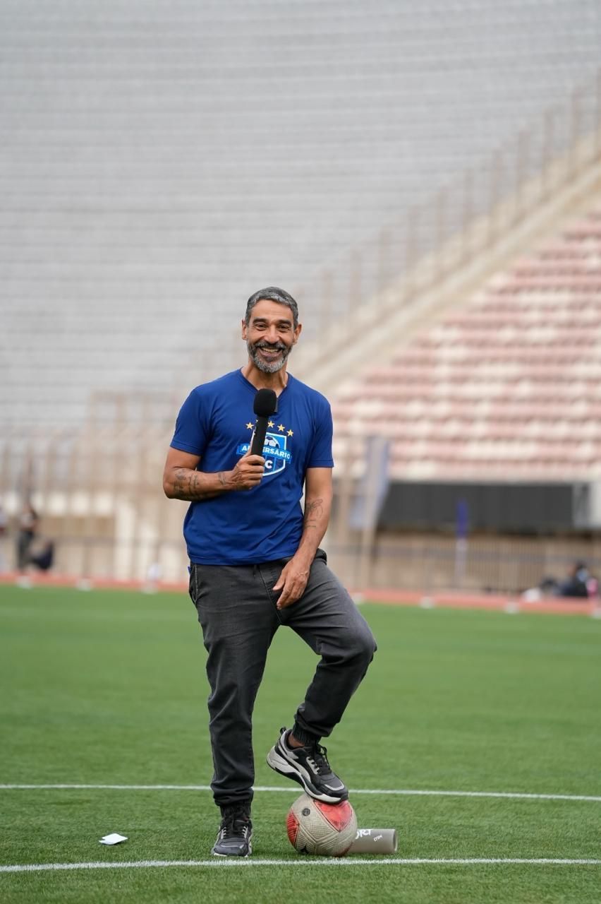Homem com microfone em pé num campo de futebol, com um pé sobre uma bola de futebol. Estádio ao fundo.