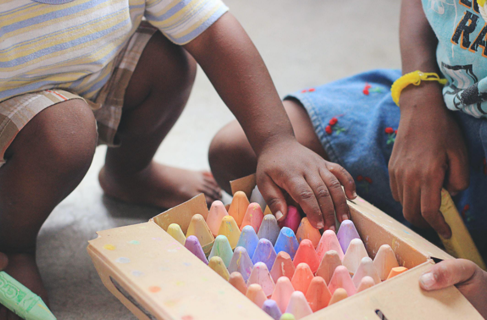 Two children are playing with a box of crayons on the floor.