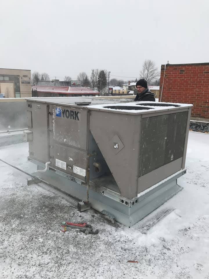 Un homme se tient à côté d’un grand climatiseur au sommet d’un toit enneigé.