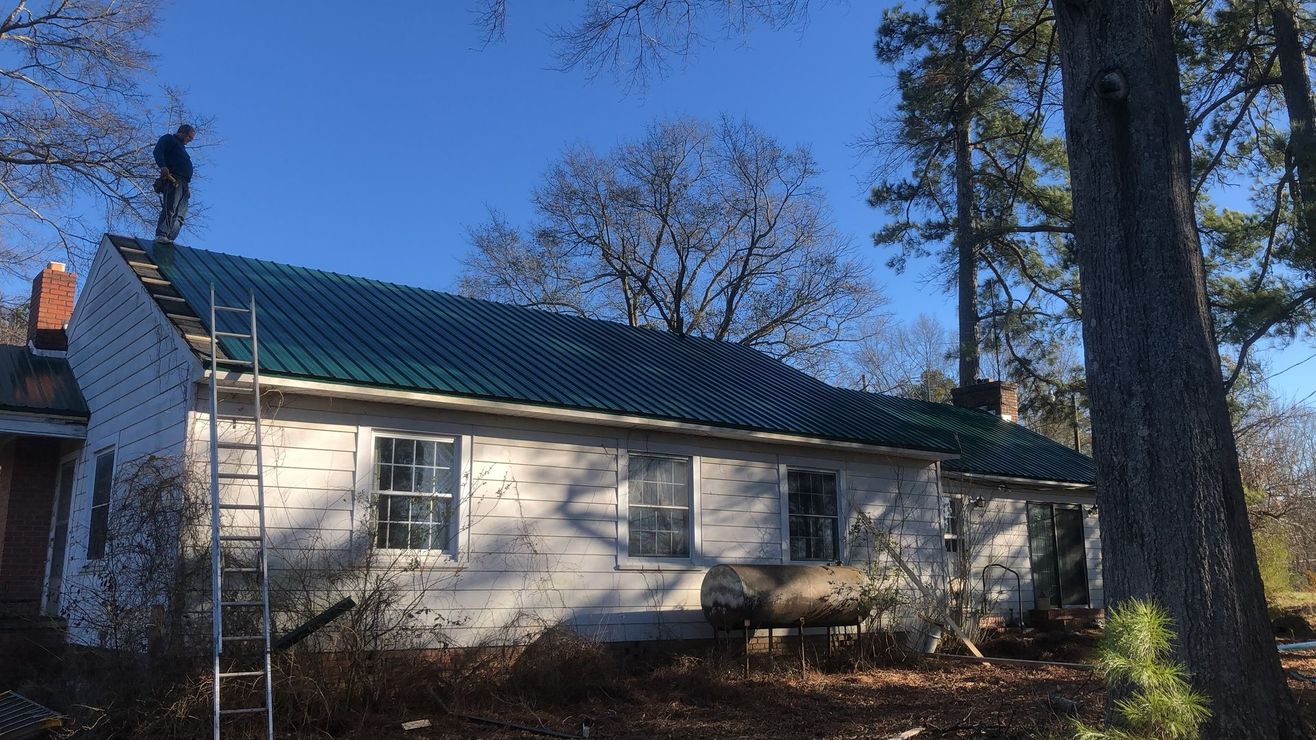A worker stands on a white house with a new green metal roof, with a ladder leaning against the side near tall trees.