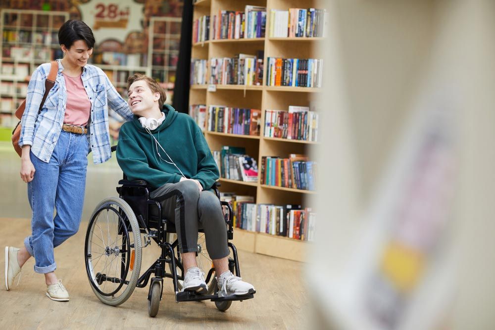 Cheerful Positive Man Disabled Student With Headphones On Neck Sitting On Wheelchair And Talking To Groupmate While They Visiting Library Together — Occupational Therapist in Kilgra, NSW