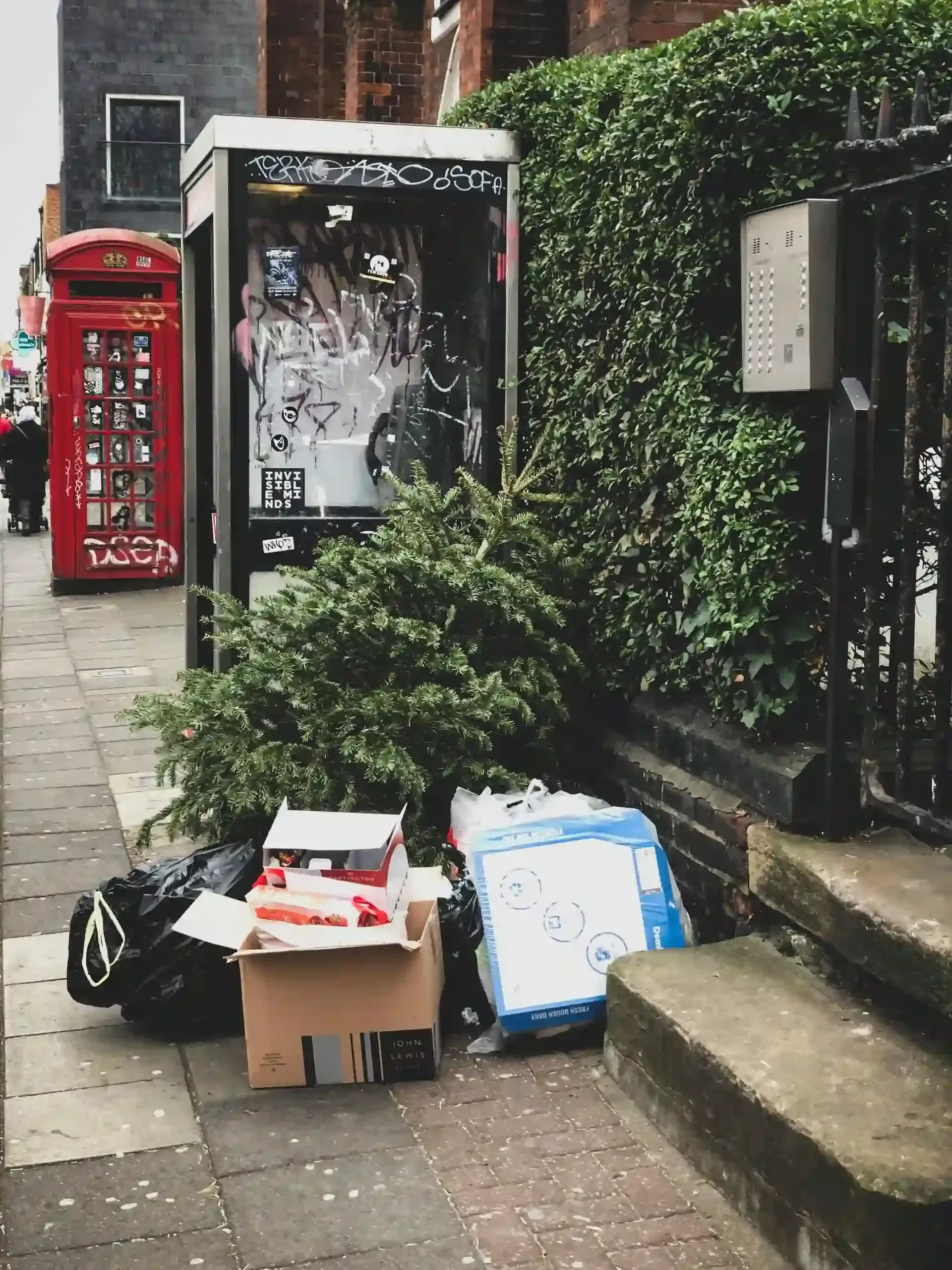 A christmas tree is sitting on the sidewalk in front of a red phone booth