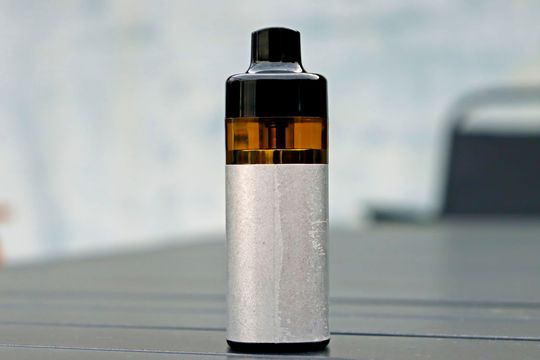 Penna per svapare, tappo nero, liquido ambrato visibile, corpo testurizzato argentato, appoggiata sulla superficie.