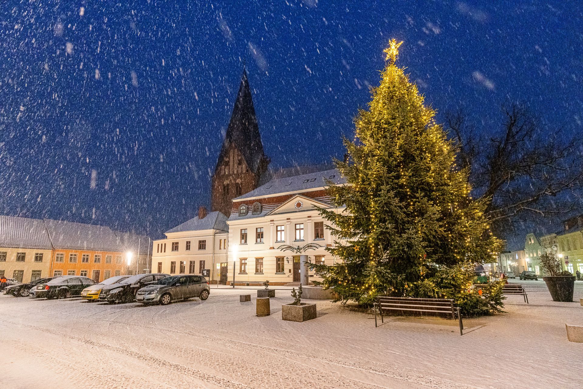 Verschneite Nachtszene mit einem geschmückten Weihnachtsbaum auf einem Marktplatz, beleuchteten Gebäuden und einer Kirche.
