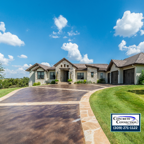 Exterior of a stone house with a brown concrete driveway, on a sunny day.