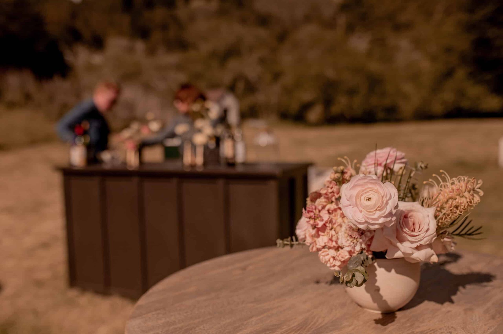 Close-up of flowers in a vase on a table, with a blurred outdoor bar and people in the background.