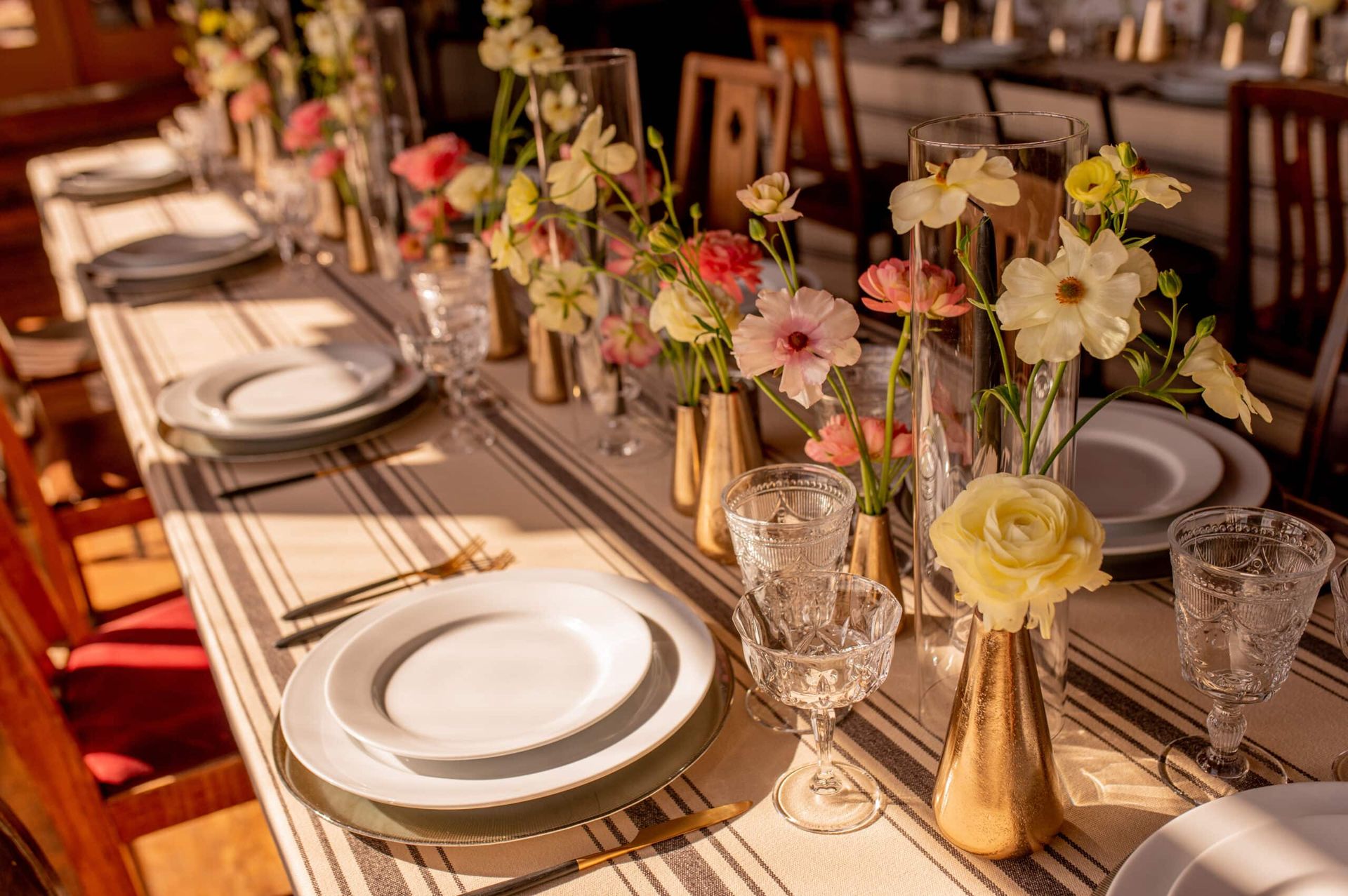 Elegant table setting with floral centerpieces, white plates, and stemware, bathed in sunlight.