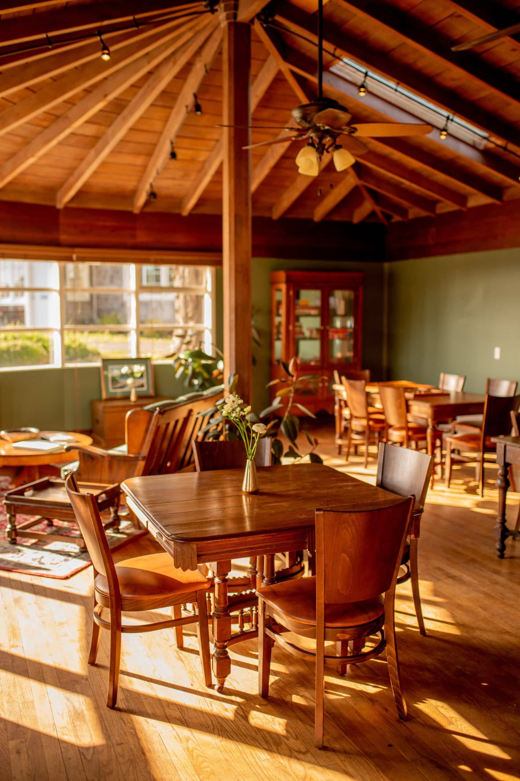 Wooden-paneled dining room with tables, chairs, and a central pillar. Sunlight streams through a window.